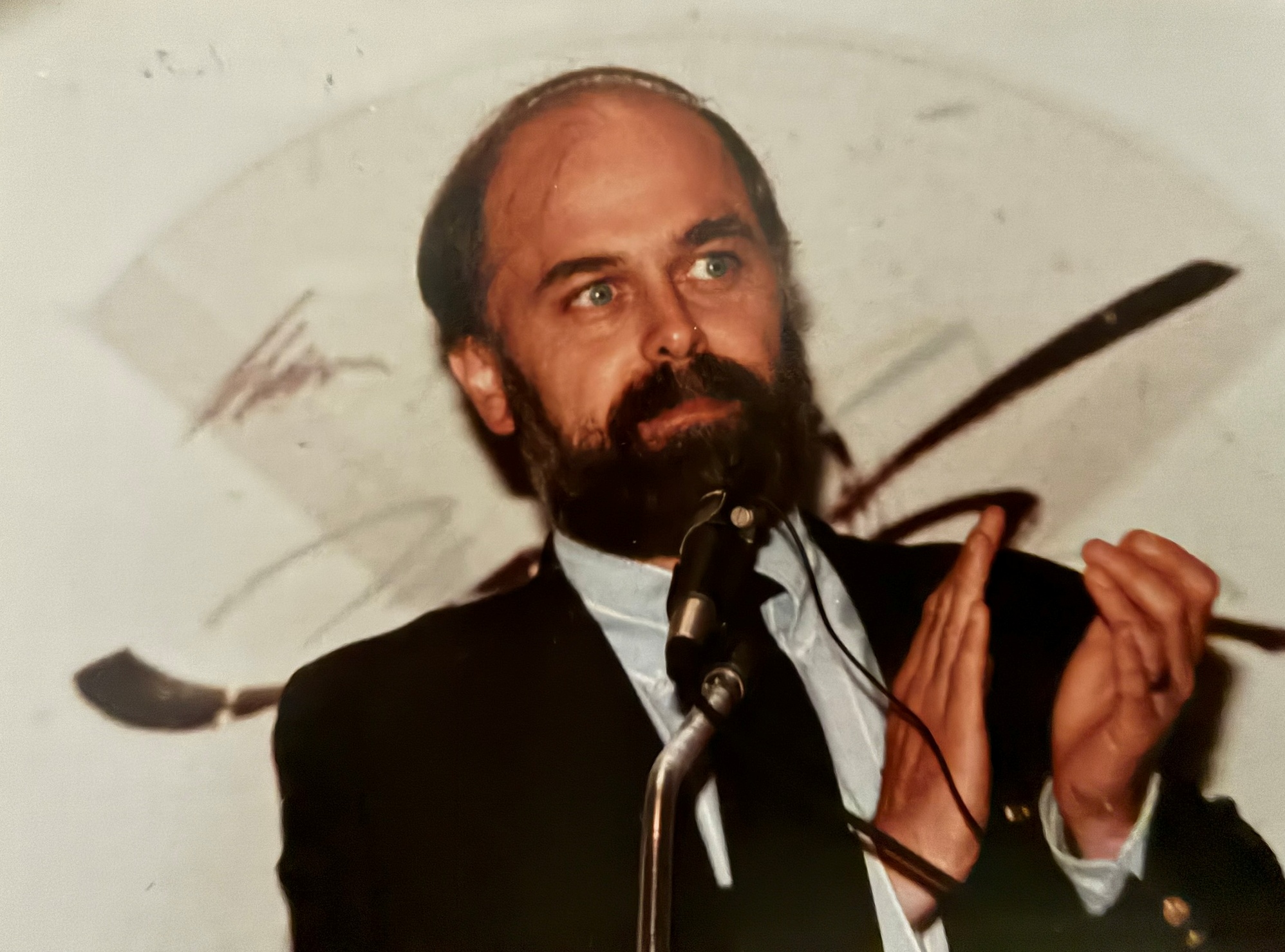 Dr. John Carey speaking at a microphone at an early SOFT event, wearing a dark jacket with dark hair and beard.