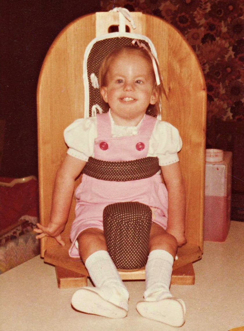 Kari Holladay at age 2, seated in an adaptive chair, smiling brightly. Kari had trisomy 18 and her referral to Dr. John Carey in 1979 led to the founding of SOFT.